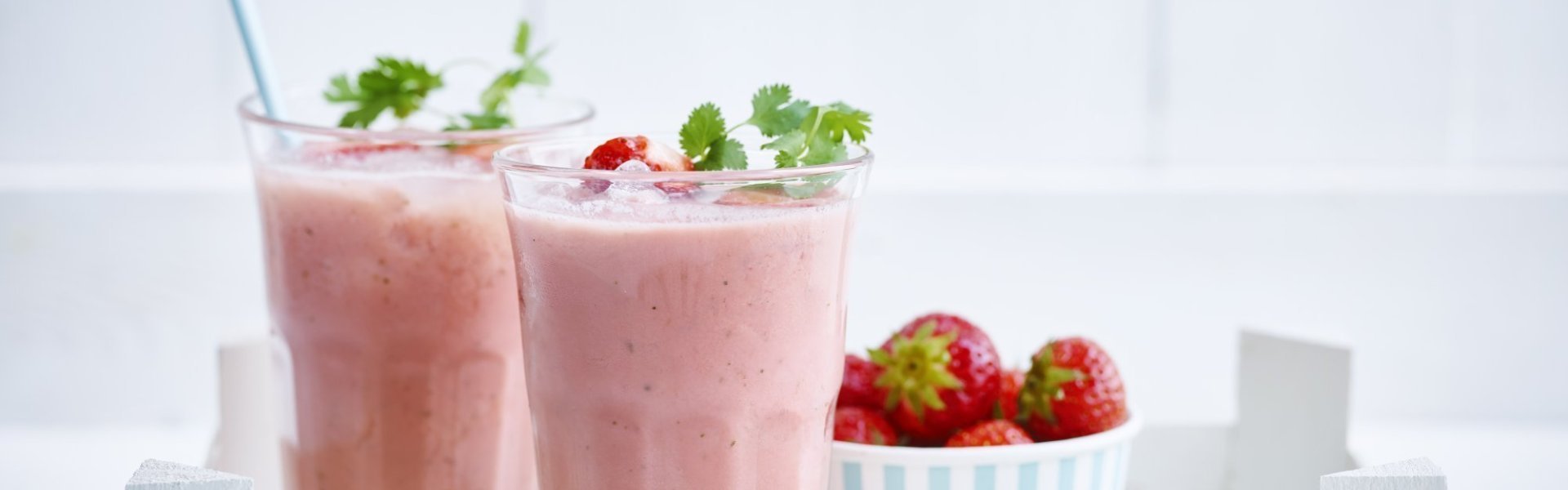 Aardbeiensmoothie 2 smoothies van aardbeien en melk. Ze staan in een houten bakje met een portie verse aardbeien er naast. De smoothies zijn afgewerkt met een takje koriander en kartonnen licht blauw rietje