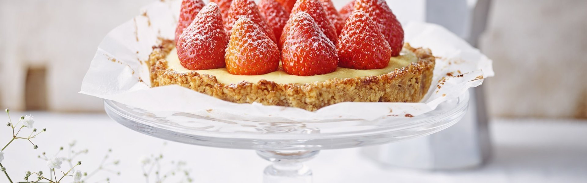 Aardbeientaart met vanillepudding en notenbodem De taart staat de stralen op een kristallen taartplateau. De verse aardbeien op de taart stelen de show. Achteraan de foto staat een kleine percolator om verse koffie te maken.