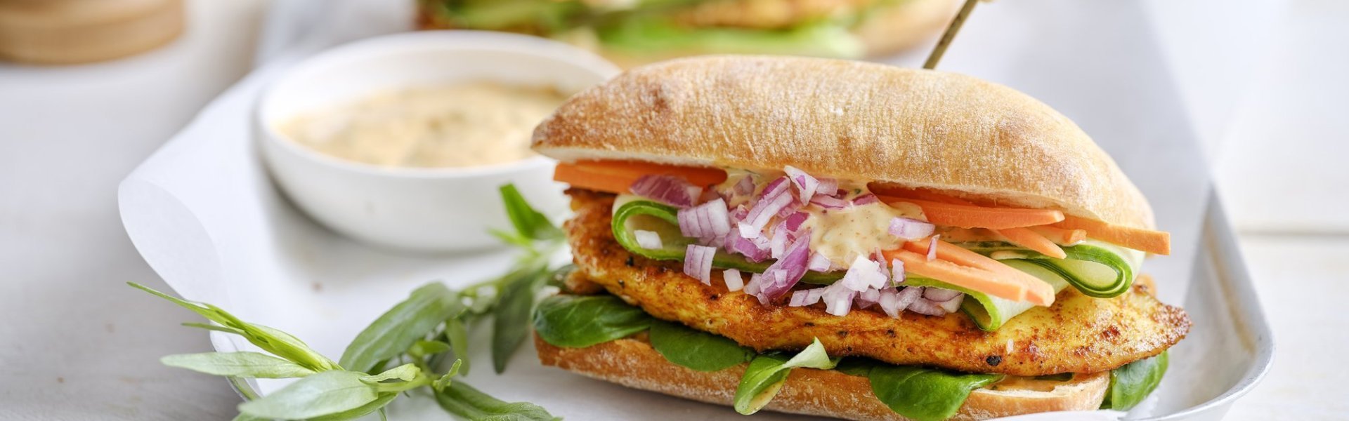 Belegd broodje met gegrilde kip Een belegd broodje ligt op een bordje. Hij is belegd met een stukje gebakken kipfilet, verse wortel, komkommer en een sausje. De resterende saus werd in een klein potje erbij geserveerd.