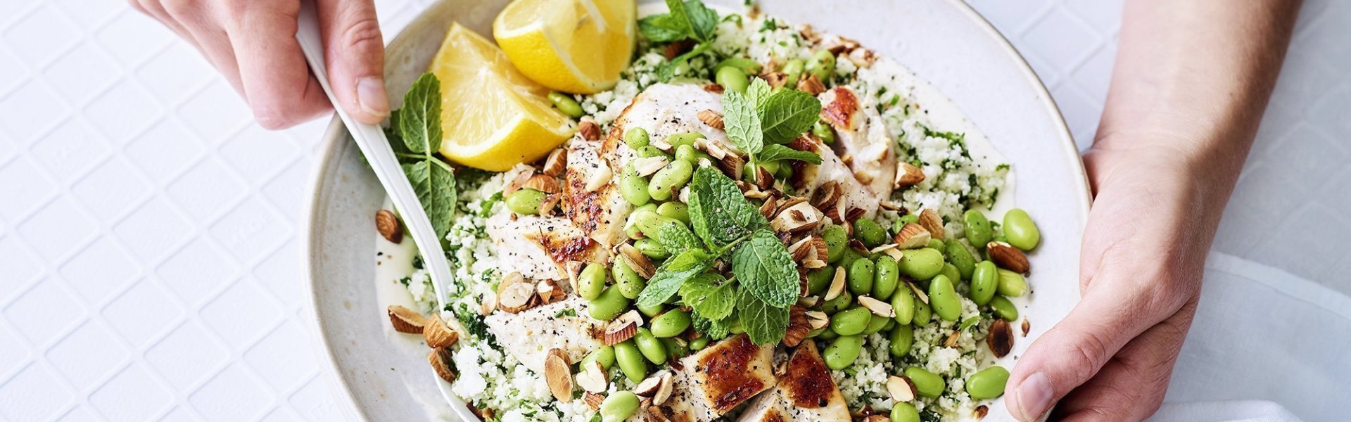 Bloemkoolrijst met kipfilet en groene groenten Bloemkoolrijst met gebakken stukjes kipfilet en veel groene groenten in een wit bord. Het bord staat op tafel met een wit tafelkleed. De vrouw die aan tafel zit schept de bloemkoolrijs om. De vrouw draagt een blauwe jeans en lichtblauwe t-shirt.