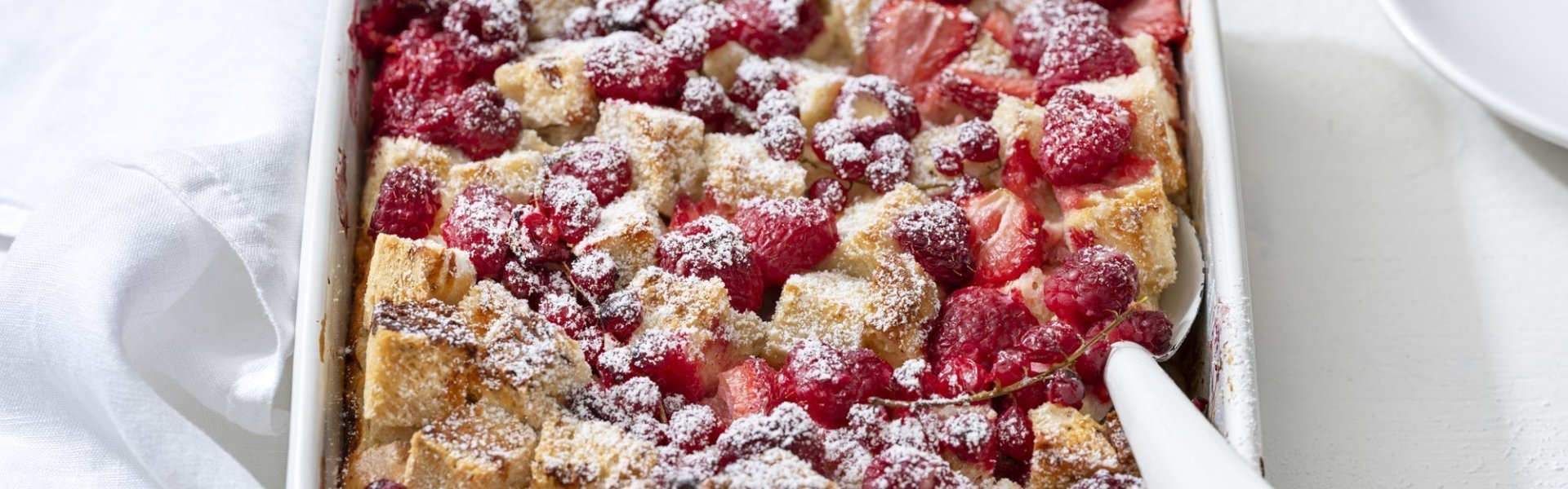 Broodpudding met rood fruit Op een witte houten tafel staat een rechthoekige ovenschotel met hierin de broodpudding klaargemaakt. Hij oogt zeer mooi door de toevoeging van het rode fruit en de afwerking met bloemsuiker. Er staan bordjes langs de ovenschotel om onmiddelijk op te scheppen en eventueel nog wat extra rood fruit te nemen.