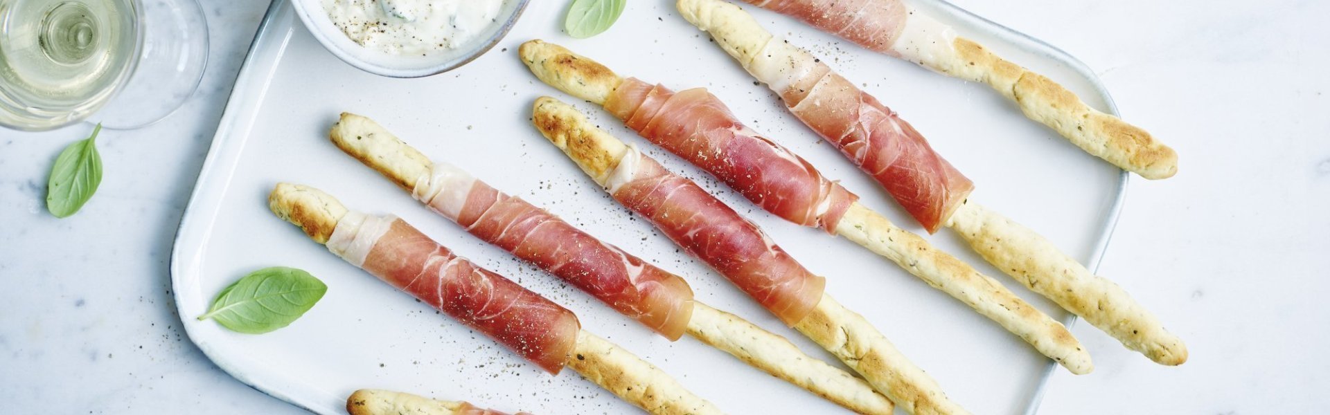 Broodstengels met gedroogde ham op een wit dienblad. Linksboven staat een potje met een dip op basis van blauwe schimmelkaas. Er staan ook twee glazen witte wijn bij en blaadjes verse basilicum ter versiering. 