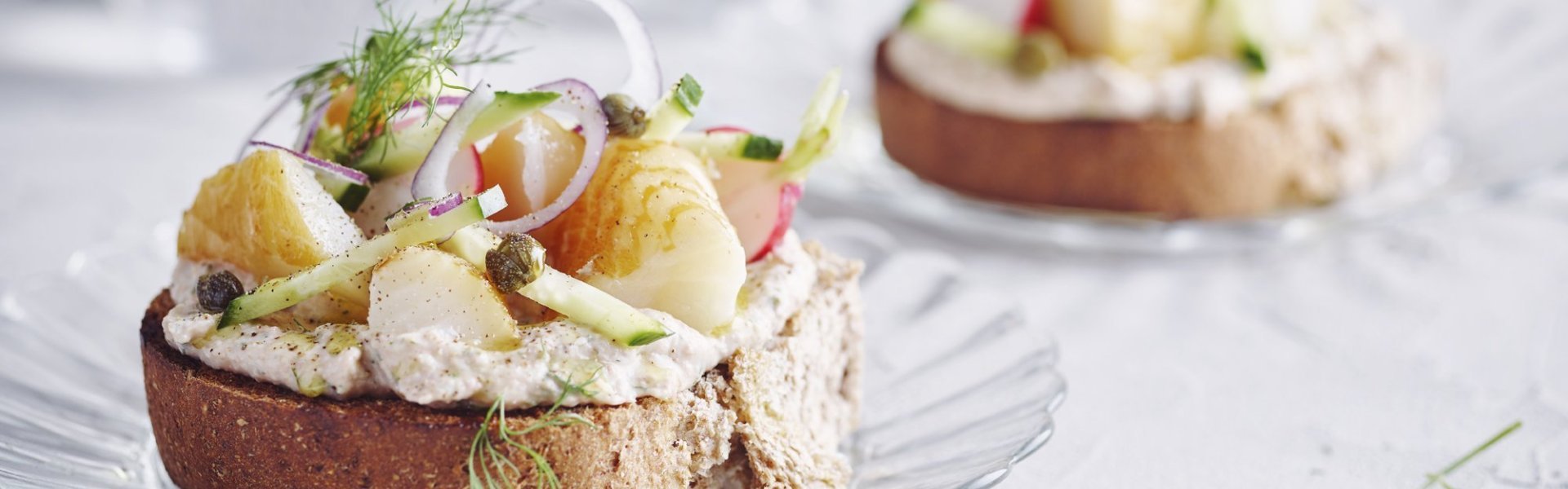 Bruschetta met bonenpuree, gerookte heilbot, radijzen en komkommer Een goedgevulde bruschetta ligt op een klein glazen bordje en staat op tafel. De bruschetta werd afgewerkt met een takje dille. Achteraan de tafel staat een kan met water en bestek om deze toast te verorberen.
