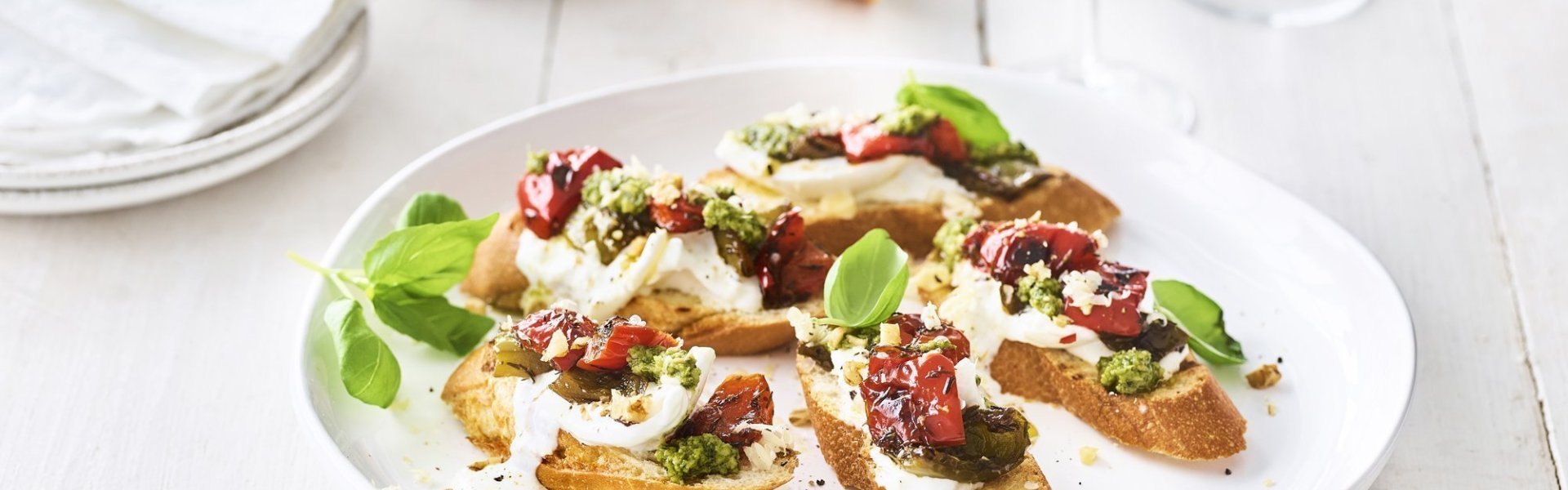 Bruschetta met paprika, burrata en pesto van basilicum met walnoten Op een houten, witte tafel staat een groot wit rond bord met hierop de bruschetta geserveerd. Ze zijn afgewerkt met extra blaadjes basilicum. Er staan ook kleine bordjes naast en de vijzel waarin de pesto is in gemaakt.