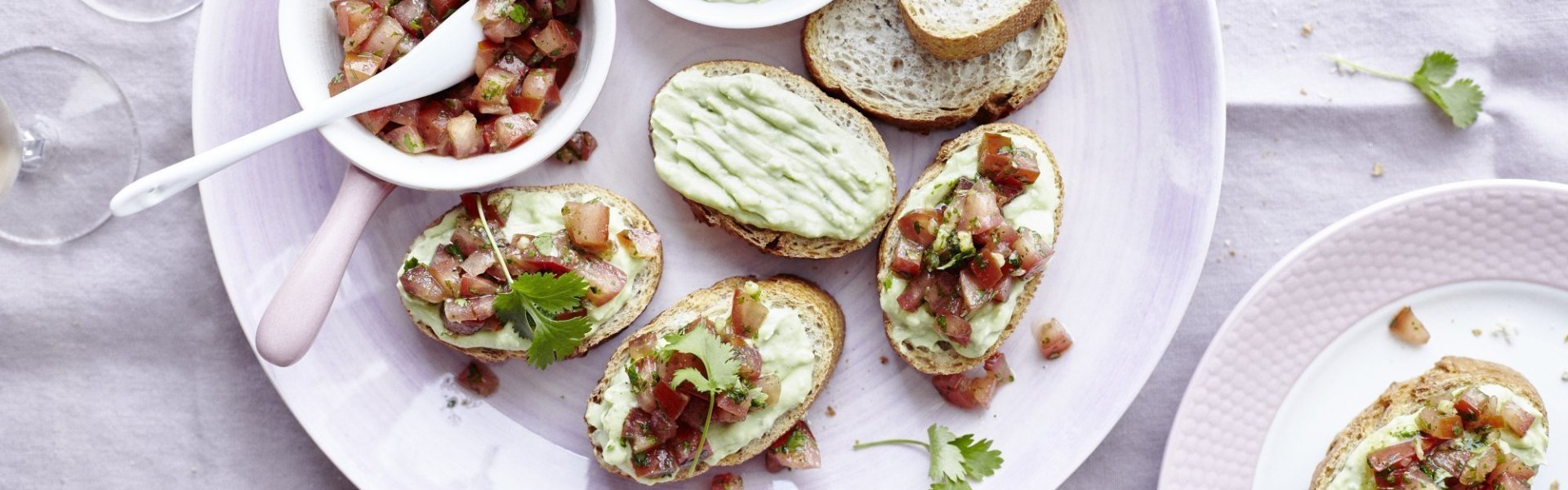 Bruschetta's liggen op een pastelroos bord, samen met een wit potje waarin de yoghurtdip zit en een wit potje waarin stukjes versgesneden tomaat en kroiander zit. De gasten smeren dus zelf hun bruschetta, waardoor de gastvrouw of -heer extra tijd aan tafel krijgt.