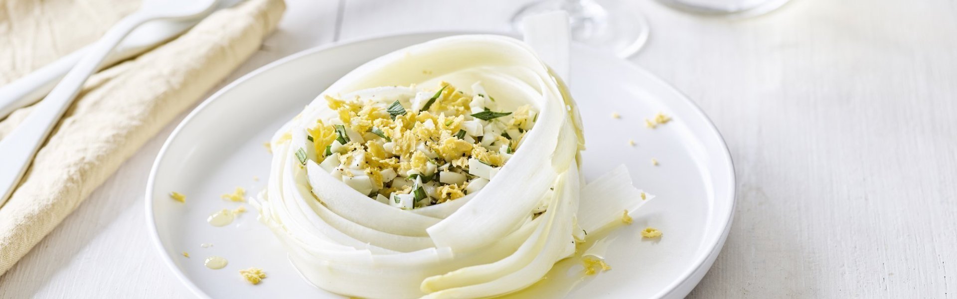 Carpaccio van witte asperge met boter en gekookte eieren De flinterdunne aspergeslierten liggen opgerold op het bord, net een vogelnestje. Hierin liggen de gehakte gekookte eieren en dragon. Er ligt bestek langs en een linnen servette.