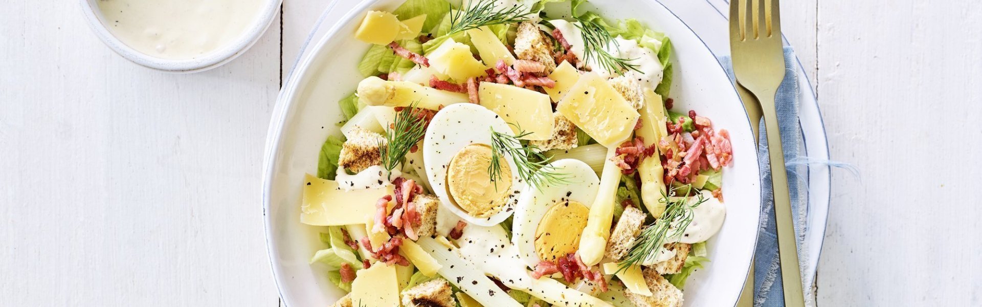 Caesar salade met asperges Een rijkgevulde salade staat klaar op tafel. Het witte bord is gevuld met sla, gekookte eitjes, stukjes asperges en kaasschilfers. Dat is de lente proeven in je bord, als je het mij vraagt!