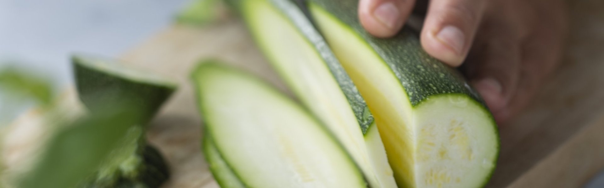 Vrouw snijdt courgette in lange repen op een houten snijplank. De vrouw heeft een licht blauwe schort aan, je ziet enkel haar midden.