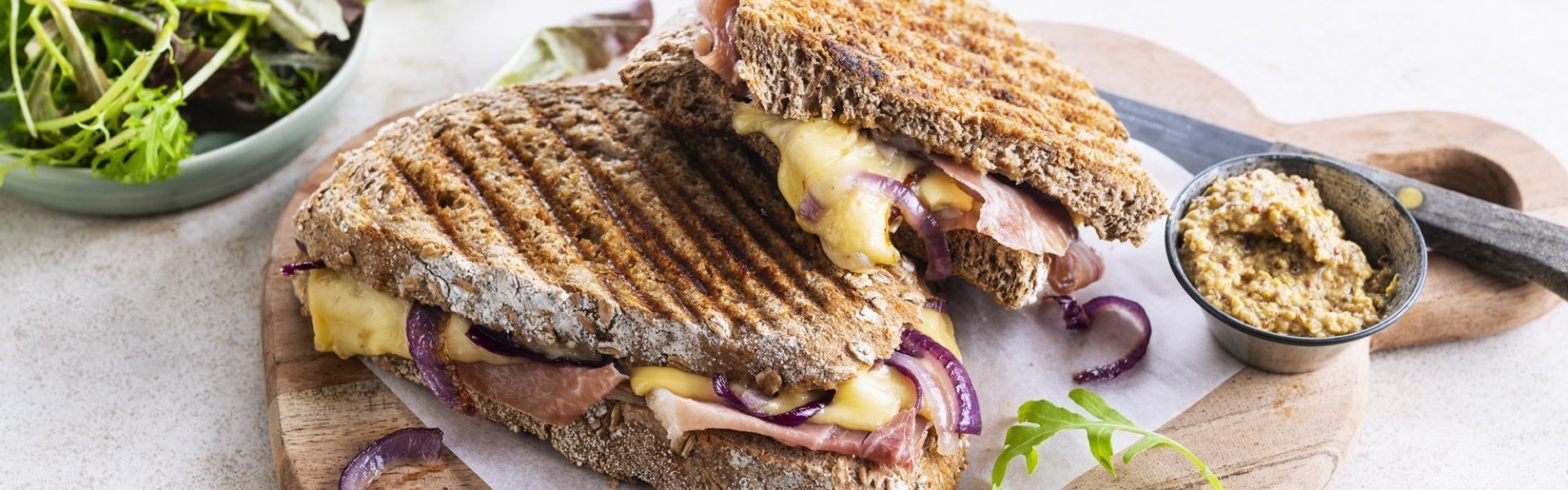 Croque met ham, rode ui en Passendale kaas De croque ligt op een houten plankje aangesneden. De kaas en rode ui zien er heerlijk uit. In de achtergrond zie je ook nog wat gemengde sla en een potje graantjesmosterd.