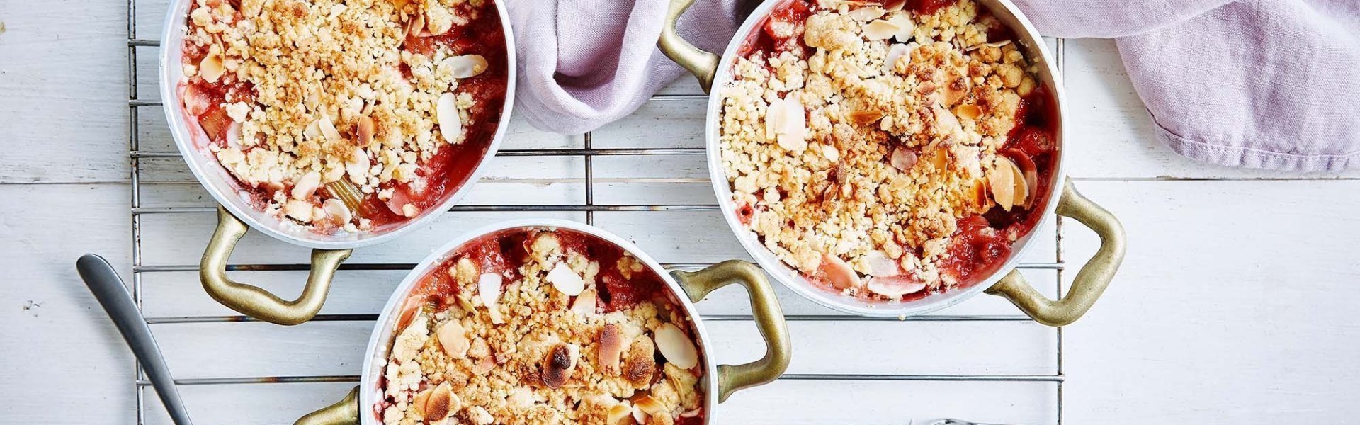 Crumble van aardbei en rabarber in kleine bronzen potjes, net uit de oven. Bovenaan de foto ligt een linnen roze servette. Alles staat op een witte houten tafel.