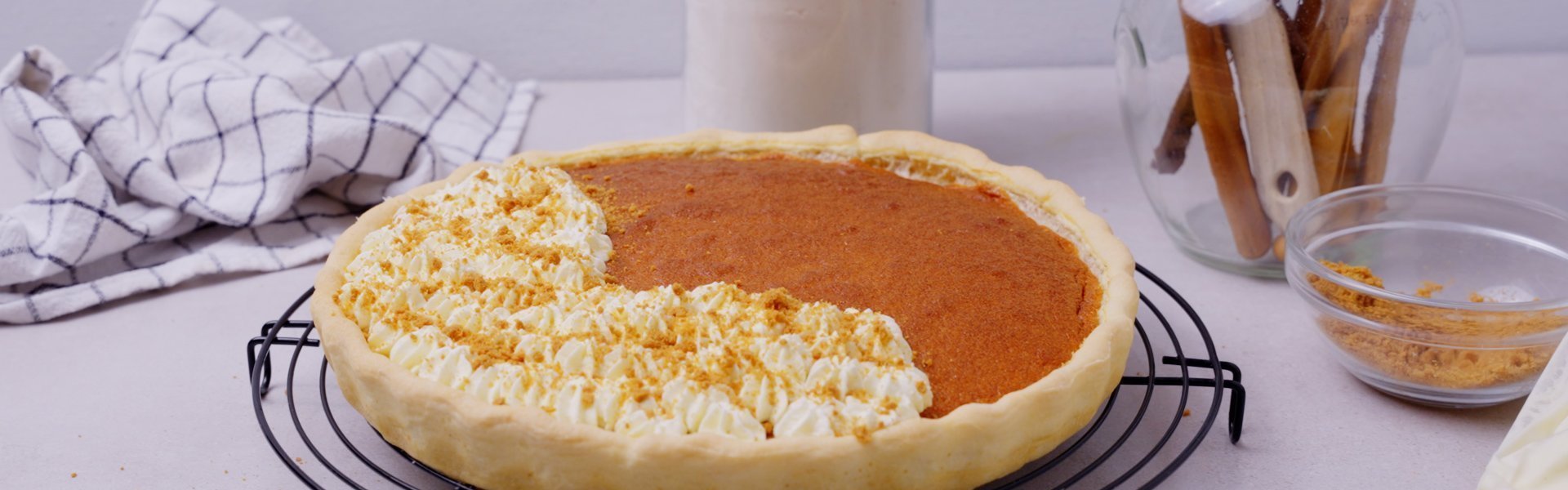 Flantaart met speculaas Een flantaart staat nog op de taartrooster, hij is half afgewerkt met toefjes slagroom en bestrooid met speculaaskruimels. Achteraan in beeld zie je de pot bloem staan en een verzamelpot met houten lepels.