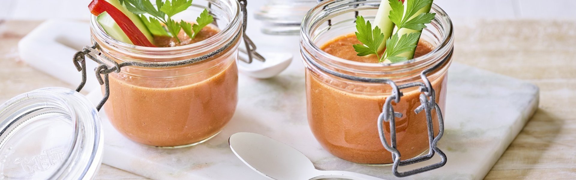 Twee bokaaltjes met gazpacho staan op een marmer plankje, een ideaal alternatief voor te weinig glazen. We gaven de gazpacho een feestlook met staafjes komkommer en paparika in het bokaaltje te laten op springen.  