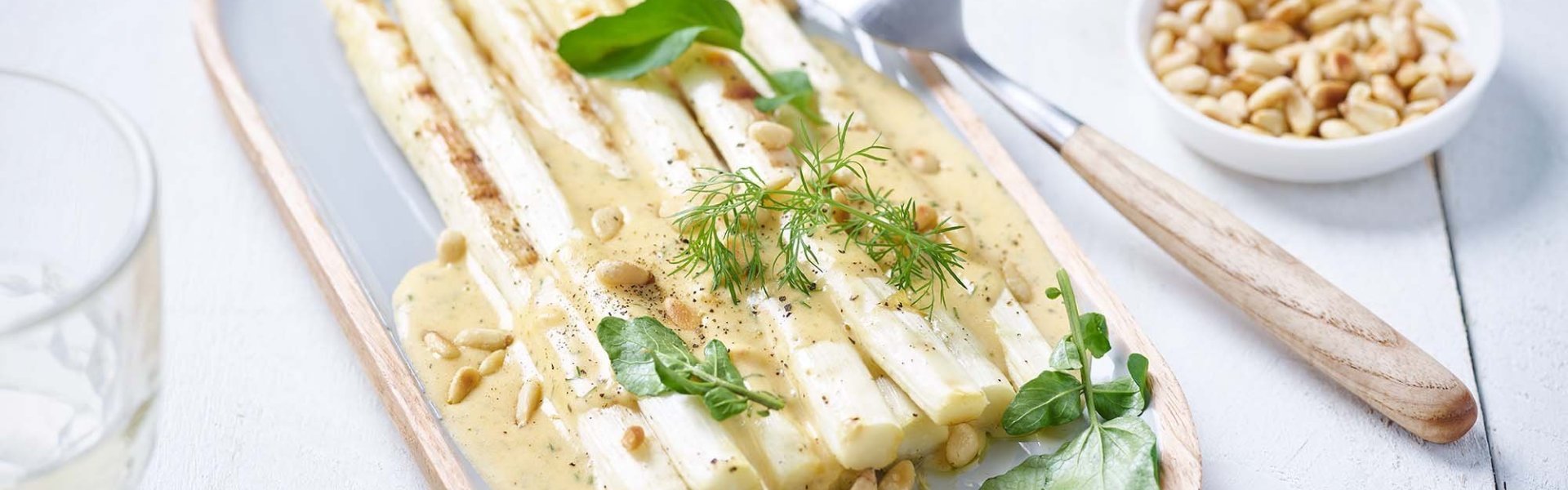 Gegrilde asperges liggen op een langwerpig ovaal bord. Ze zijn de helft overgoten met mousselinesaus en verse groene tuinkruiden. Er staat ook een potje met gegrilde noten langs het bordje.