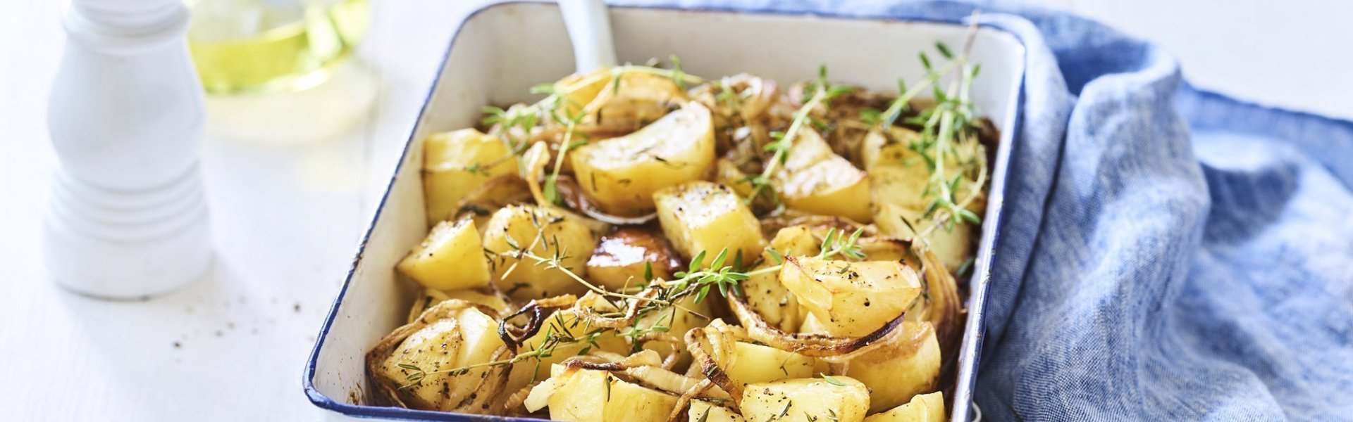 Geroosterde aardappelen met balsamico Een witte ovenschaal met donkerblauw randje staat op een keukenonderlegger op een witte houten tafel. Er staat een witte pepermolen en een glazen flesje met olijfolie langs. De verse tijm zorgt voor extra kleur bij de geroosterde aardappelen, er ligt ook nog een blauwe linnen servette langs de ovenschotel.