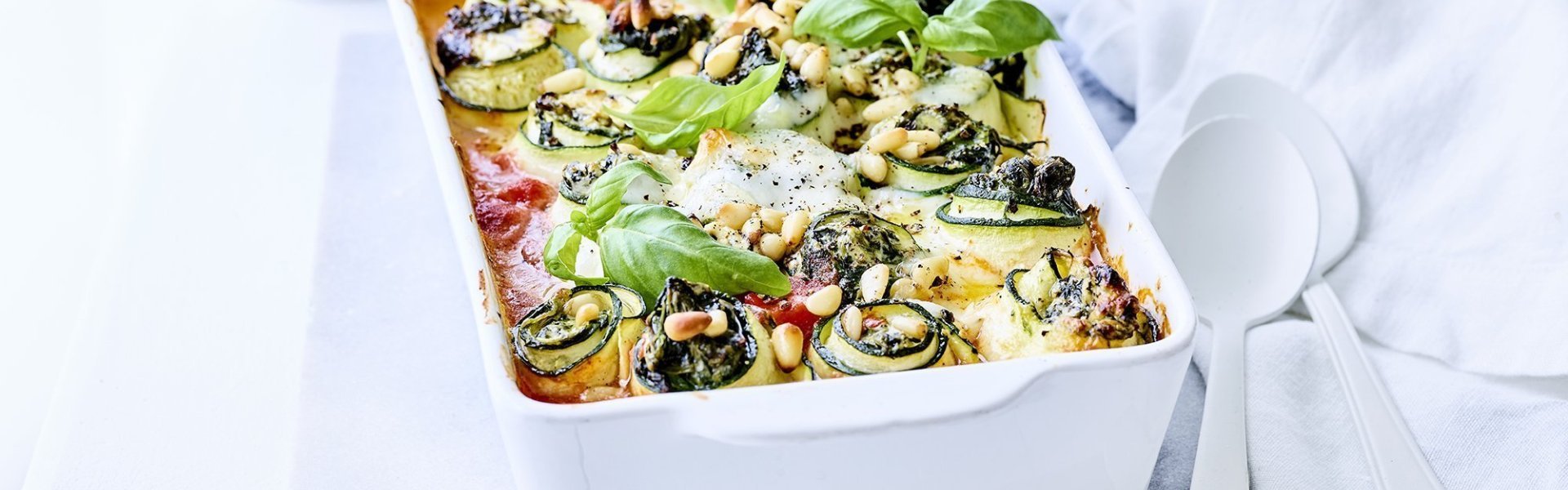 Witte ovenschaal gevuld met courgette. In de courgetterolletjes zit ricottakaas, spinazie en Belgische geraspte kaas. De ovenschotel is gevuld met tomatensaus en afgewerkt met verse basilicum. 