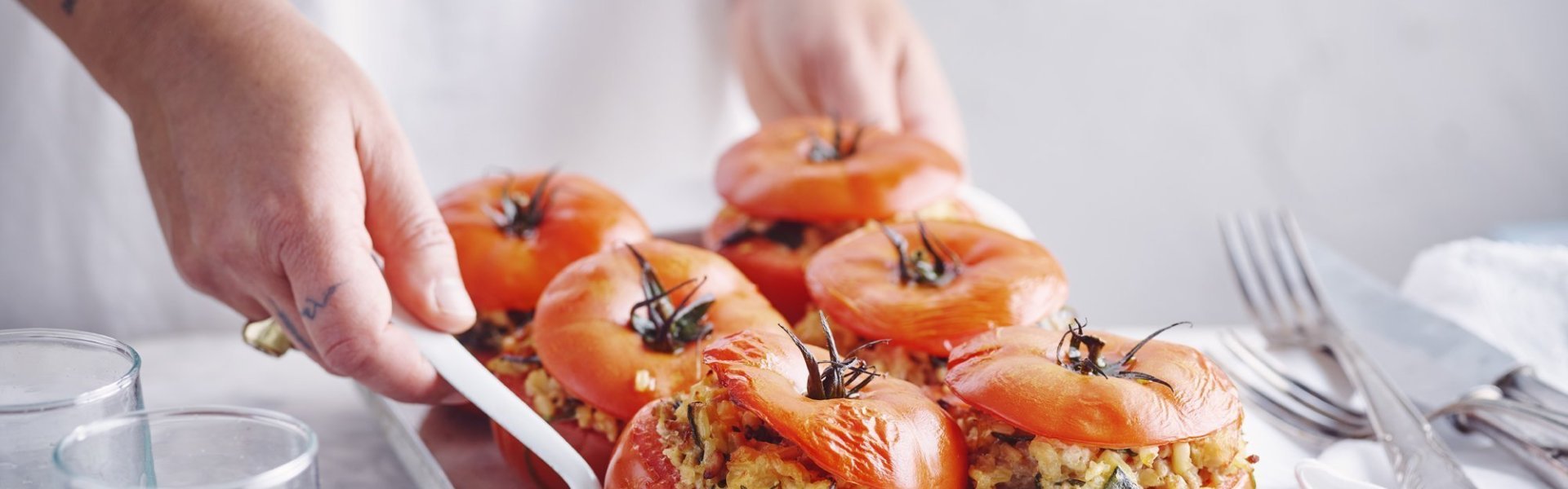 Zes gevulde rode tomaten in een ovenschaal zijn aan tafel gebracht door een vrouw in witte t-shirt. Ze schept alvast eentje op voor zichzelf.