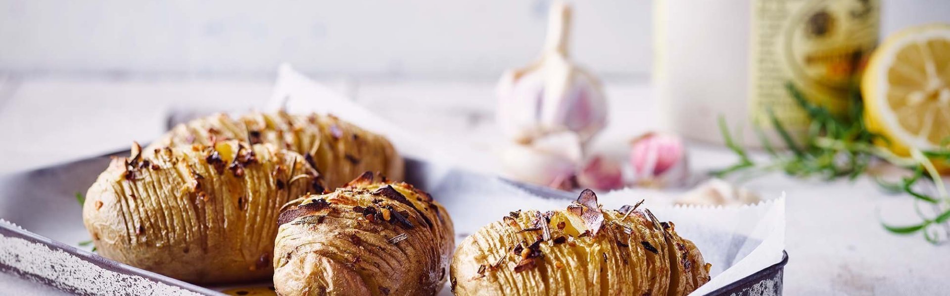 Hasselback aardappel met pittige kruiden Klaargemaakte hasselback potatoes met pittige kruiden. Ze liggen op een grijze ovenschaal op de keukentafel. Achteraan het beeld zie je hete pepers, look, rozemarijn en een halve citroen liggen.