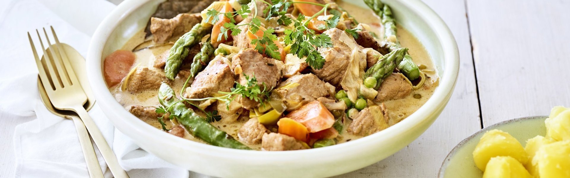 Kalfsfricassee met lentegroenten Deze fricassee straalt een en al lente uit! Hij is klaargemaakt met jonge worteltjes, erwtjes en aspergepunten. Er ligt gouden bestek langs en een portie gekookte aardappelen.