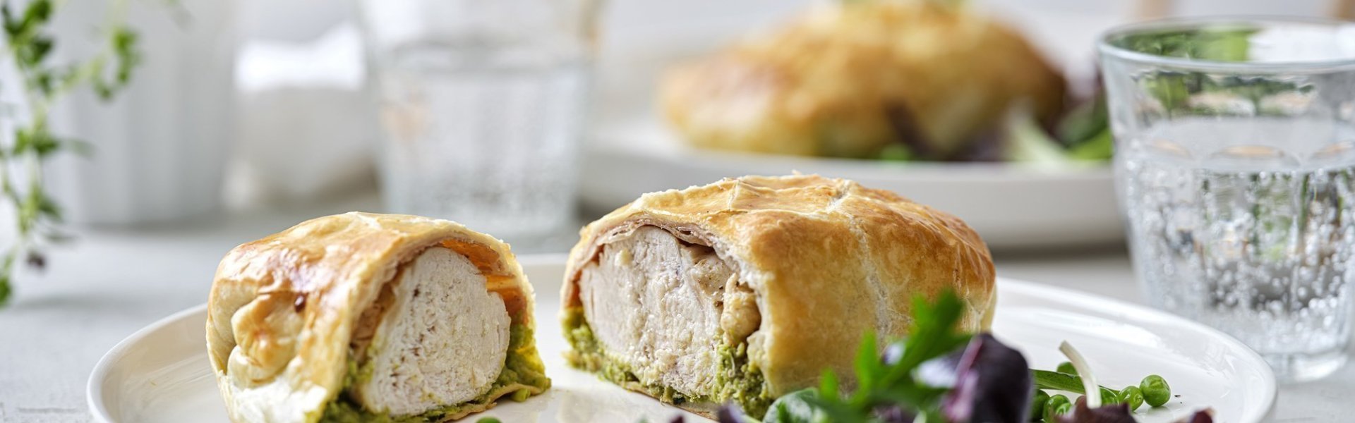 Kip wellington met gedroogde ham en erwtjespuree met munt Een mini wellington van kipfilet ligt opengesneden op een wit bord. Hierin zie je mooi een laagje erwtjespuree onder het witte vlees in het bladerdeeg pakje. Er is nog wat gemengde sla en erwtjes toegevoegd op het bord.