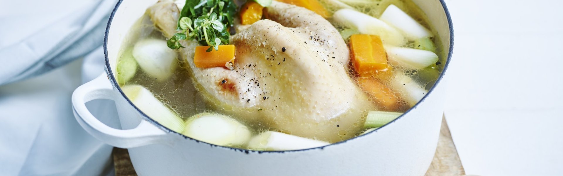 Kippenbouillon Bouillong van kip met verse groentjes erbij. De kookpot in emaille staat op een houten snijplank met verse tijm erbij en een houten lepel langs.