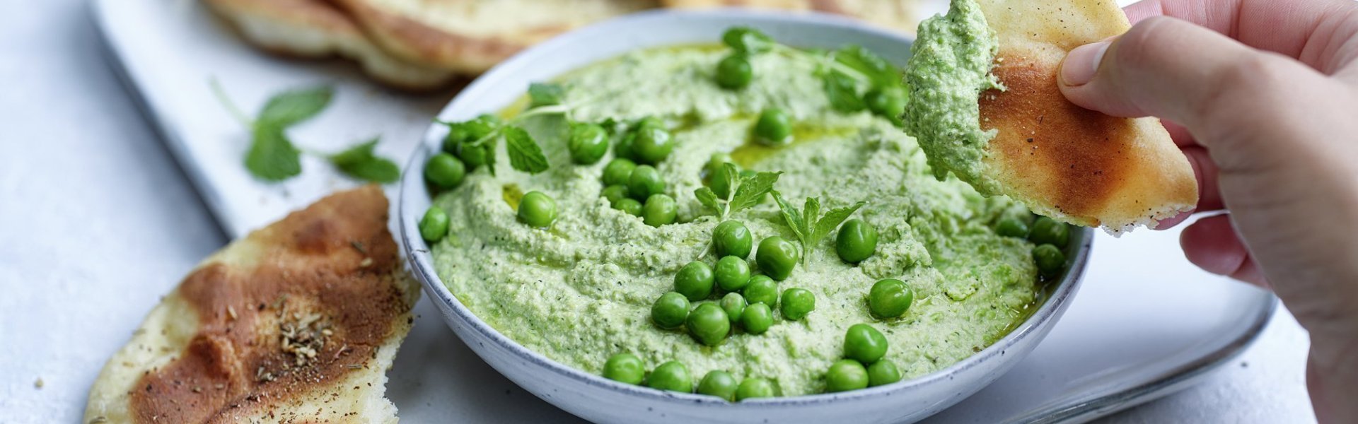 Krokant platbrood met erwtendip Krokant gebakken platbrood ligt op een serveerschaaltje waarbij een dip van erwten en roomkaas in geserveerd. De dip is gemixt en heeft een mooie groene kleur, om af te werken zijn er nog enkele verse erwtjes bovenop gestrooid. Een hand neem alvast een stukje platbrood om te dippen in gezonde groentedip.