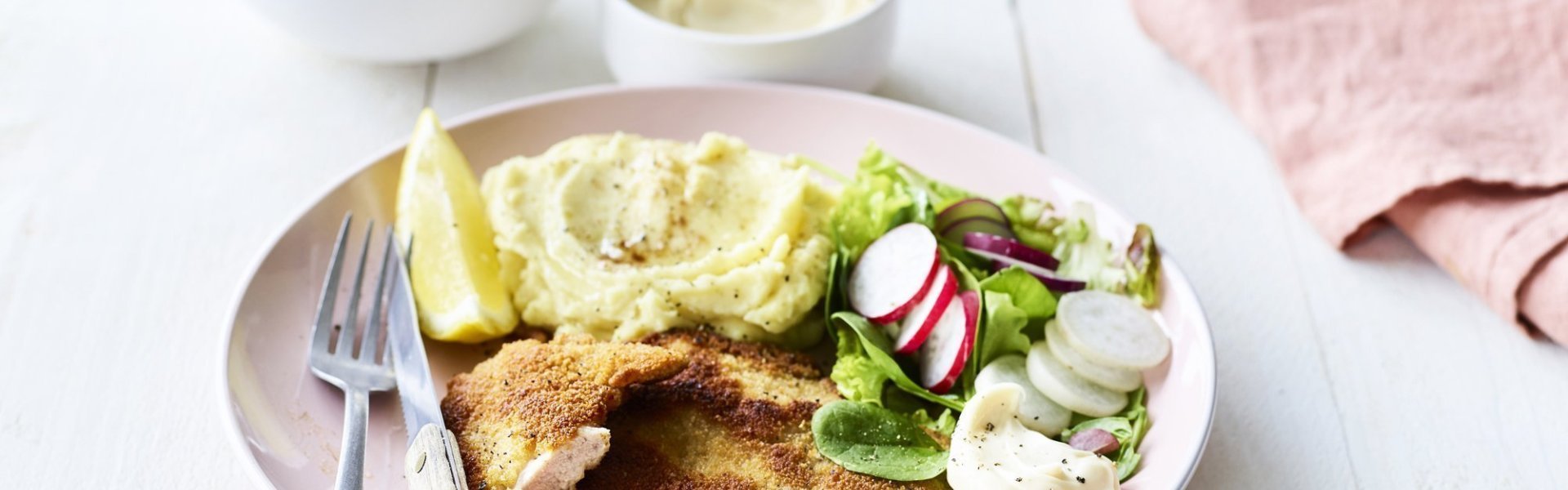 Shnitzel met zelfgemaakte mayonaise Een homemade shnitzel ligt op een pastelroze bord, samen met aardappelpuree, verse sla en zelfgemaakte mayonaise. Er is nog wat extra salade in een wit potje en mayonaise voor extra lekkers op te scheppen.