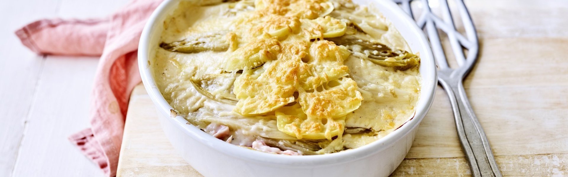 Er staat een ovenschotel op een houten plank, op de tafel. Er ligt een lepel naar de ovenschotel die gevuld is met een lasagne van witloof, ham, kaas en aardappelen. Er ligt ook een coraalroze servette bij.