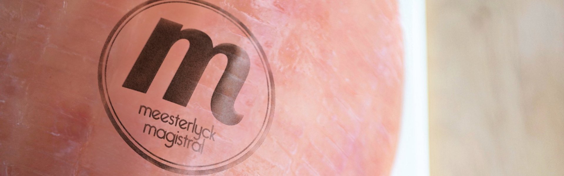 Meesterlyck-ham Volledige ham op witte porseleinen snijplank, geplaatst op een houten aanrecht. Het Meesterlycklogo is de gekookte ham gebrand.