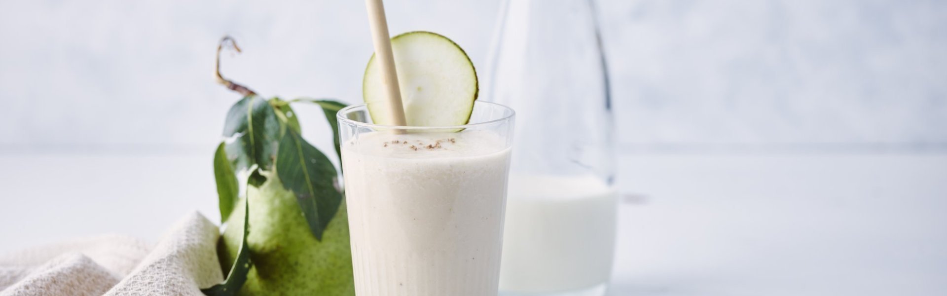 Een milkshake op basis van karnemelk, peer en banaan. Hij is geschonken in een hoog glas met rietje en stukje peer. Links van het glas ligt een verse peer en linnen servette. Rechts ligt de honinglepel op tafel.