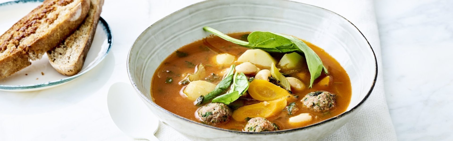 Een minestrone soep met gehaktballetjes en stukken groentjes in licht grijs diep bord. Witte lepel en vers getoast brood ligt langs het bord op de tafel. Klaar om aan te vallen. 