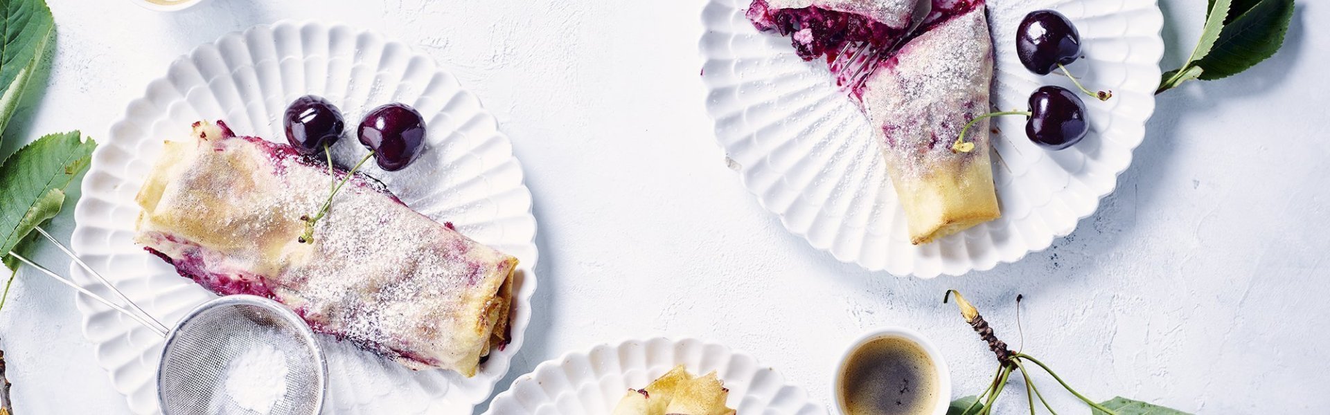 Ministrudel van kersen en platte kaas Een zomerse versie van onze appelstrudel, deze keer met kersen en plattekaas. Er staan er verschillende op tafel gepresenteerd met een kopje koffie, klaar om op te eten.