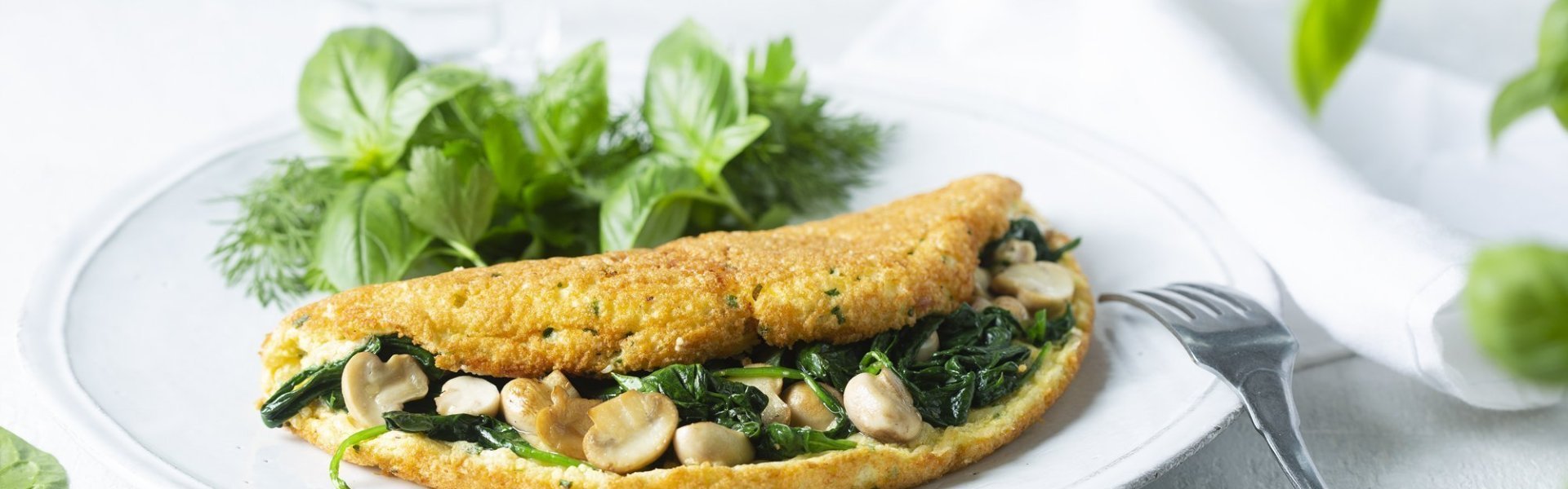Omeletsoufflé met champignons, spinazie en kaas Een heerlijke dichtgevouwde omeletsoufflé gevuld met champignons, spinazie en kaas ligt op een wit bord. Het gerechtje werd afgewerkt met een slaatje van verse kruiden, denk maar aan basilicum en dille.
