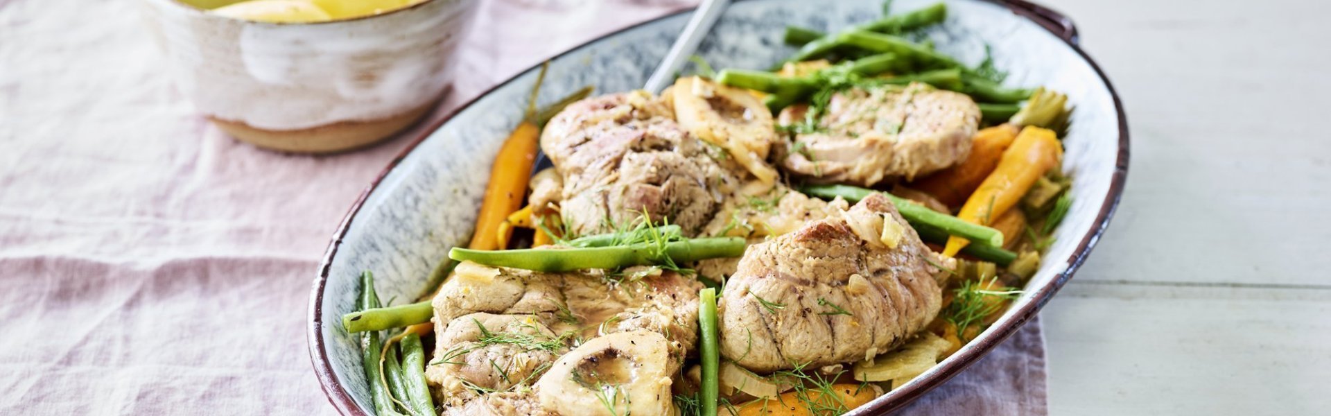 De kalfsschenkels liggen in een ovenschotel met groene boontjes en oranje wortels erbij. Dit zorgt voor een mooie variant op de klassieke ossobuco.