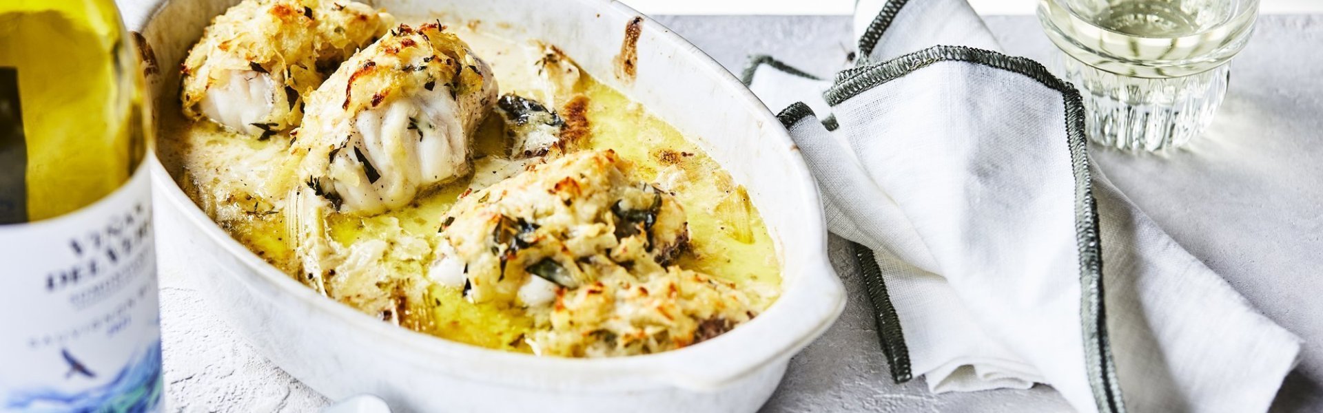 Ovenschotel met zeeduivel Een ovenschotel staat klaar op tafel om meteen te beginnen. De schotel is gevuld met stukken zeeduivel en prei in een mosterdsausje. Er ligt een krokant korstje op van geraspte aardappel en kaas. Er staat een flesje wijn langs en een witte scheplepel.