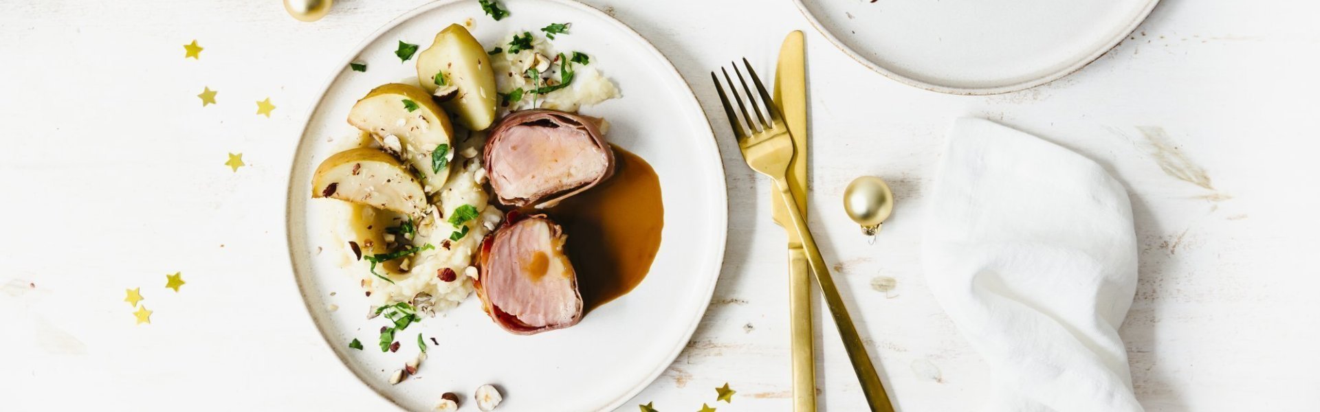 Puree van aardpeer met hazelnoot, varkenshaasje en ovenappeltjes Feestelijk hoofdgerecht van varkenshaasje met een puree van aardpeer en ovenappeltjes. Goud bestek en gouden minikerstballetjes liggen langs het bord en geeft instant een Kerslook.