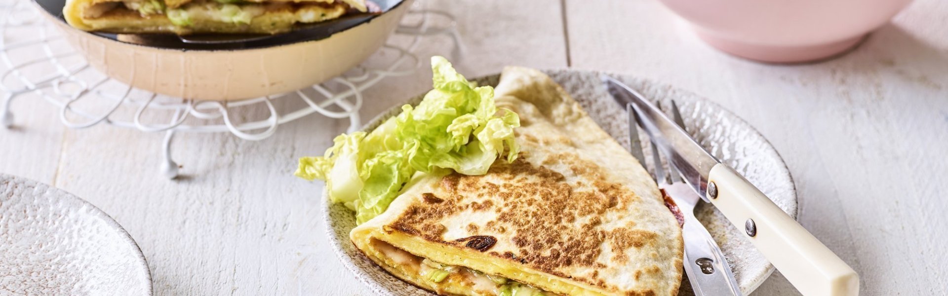 Quesadilla met een omelet en Passendale kaas Een krokante tortilla ligt in een bordje klaar om aan te vallen. Hij is gevuld met een omelet, passendale kaas, gandaham en lente-ui. De tweede quesadilla ligt nog in de pan en staat ook op de tafel.