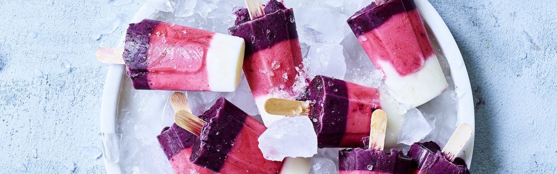 Zelfgemaakte raketijsjes met heerlijk Belgisch zomerfruit. Denk maar aan aardbeien en blauwe bessen. De homemade ijsjes liggen op een schaal gevuld met ijsblokjes. 