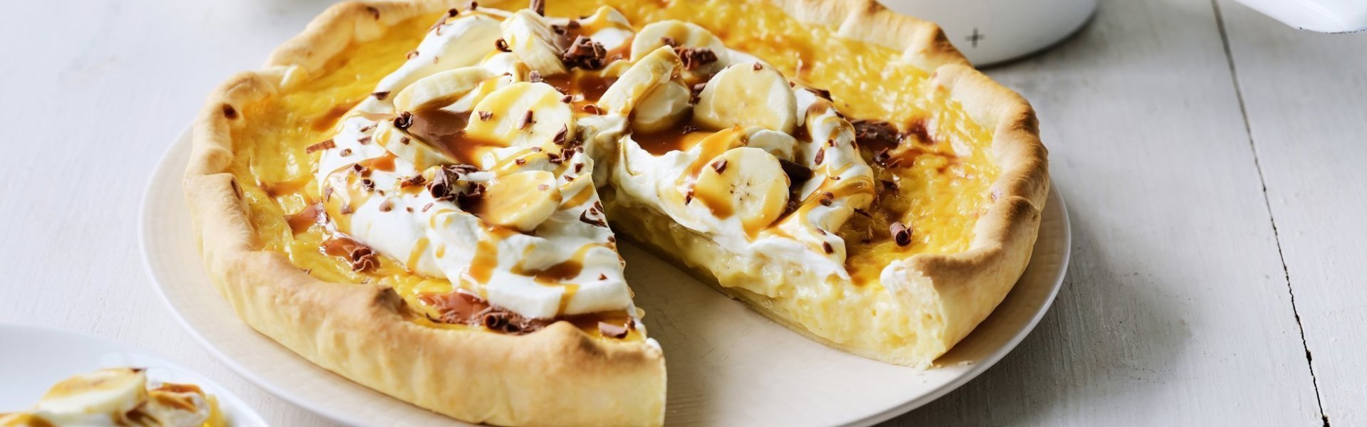 Rijsttaart met slagroom, banaan en gezouten karamel Op een witte houten tafel staat een klaargemaakte rijsttaart, hij is afgewerkt met vers opgeklopte slagroom, stukjes banaan en vloeibare karamel. Er is al een stukje aangesneden. Verder uit beeld staat het steelpannetje waarin de karamelsaus is klaargemaakt.