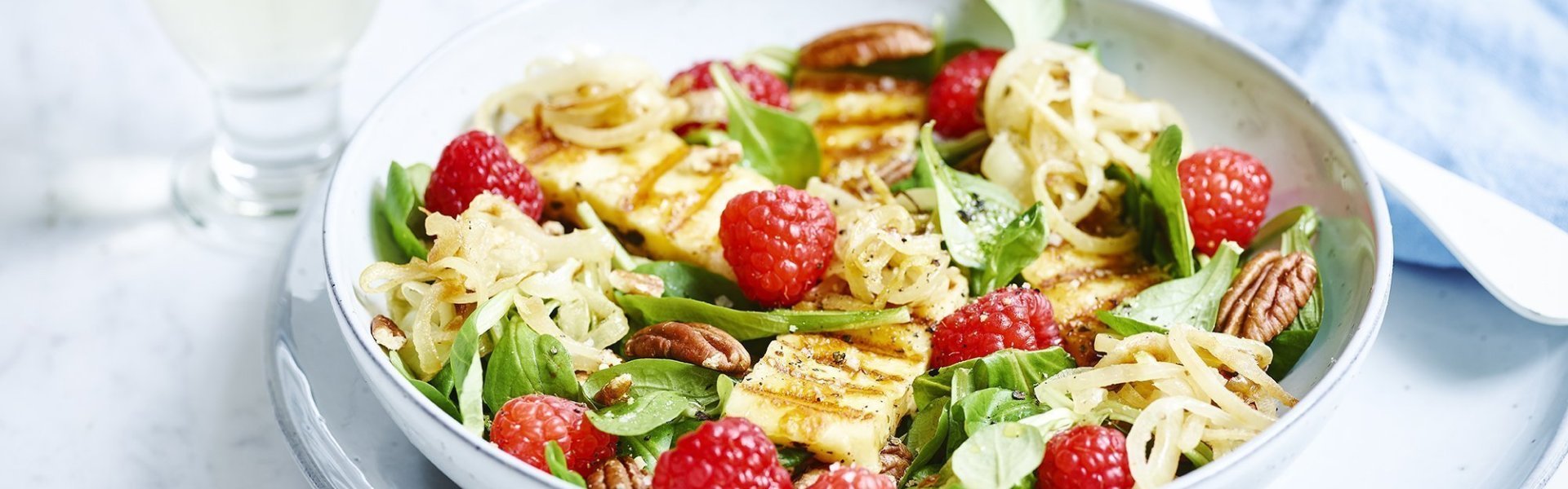 Een zomerse salade met gegrilde Berloumi kaas, dit is een Belgische grillkaas, en zomerzoete frambozen. De salade is gepresenteerd in een diep licht blauw bord, linksboven staat een glaasje witte wijn. Recht zie je wit bestek liggen op een blauwe linnen servette.