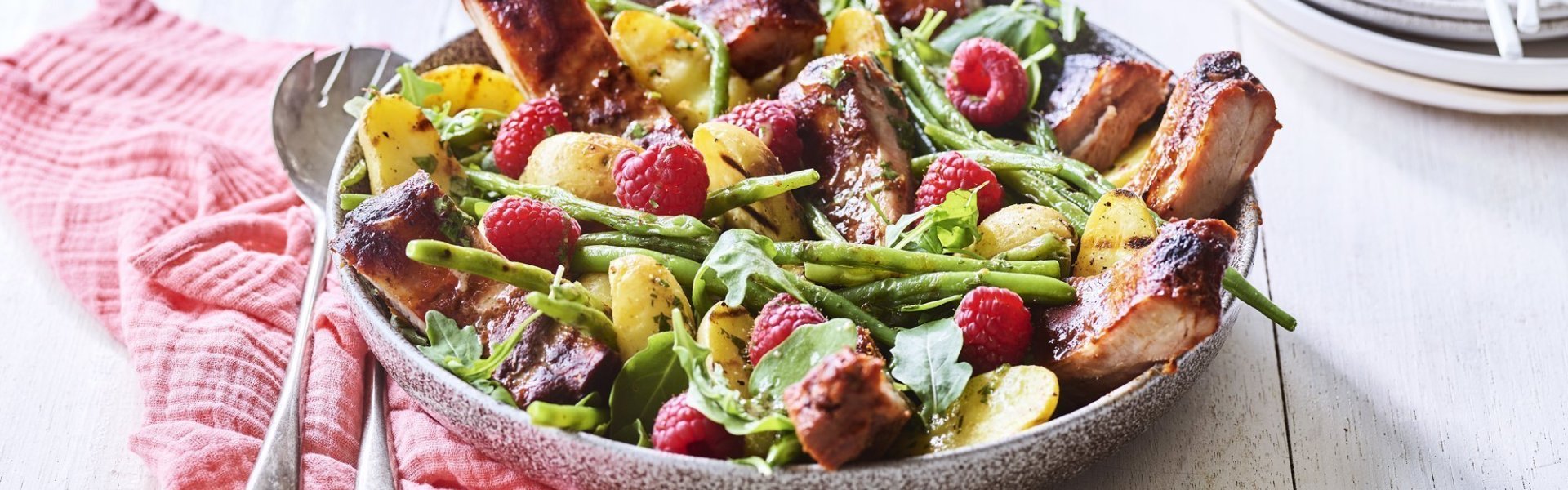 Salade van krieltjes, boontjes, frambozen en ribbetjes Een grote serveerschaal is gevuld met de aardappelsalade. Je ziet de framboosjes, boontjes en ribbetjes in de salade zitten. Er staat een torentje bordjes klaar met bestek om de salade te verdelen en op te eten.