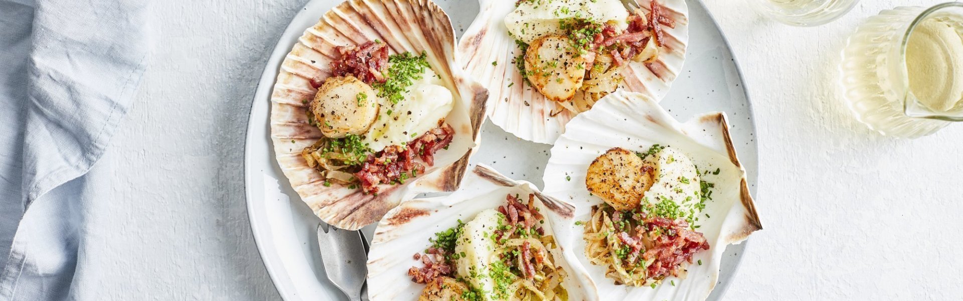 Sint-jakobsvrucht met bloemkoolmousse, witloof en spekjes Sint-jakobsvruchten op een groot bord, opgediend in hun eigen Sint-jakobsschelp met bloemkoolmousse, witloof en spekjes. Glaasjes wijn en bestek ligt mee naast het bord, klaar om het feestmaal aan te vallen.