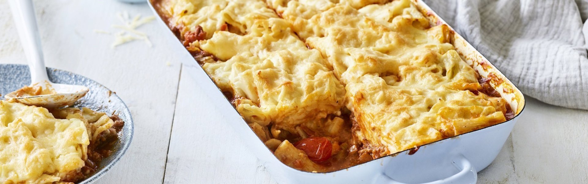 Slowcooked mac & cheese lasagne Een lichtblauwe ovenschotel staat op een houten witte tafel. De ovenschotel is gevuld met een mac & cheese én een tomatensaus, waardoor hij eerder weg heeft van een lasagne. Er is al 1 portie uitgeschept en wil al beginnen aan de volgende. Laat het niet koud worden.