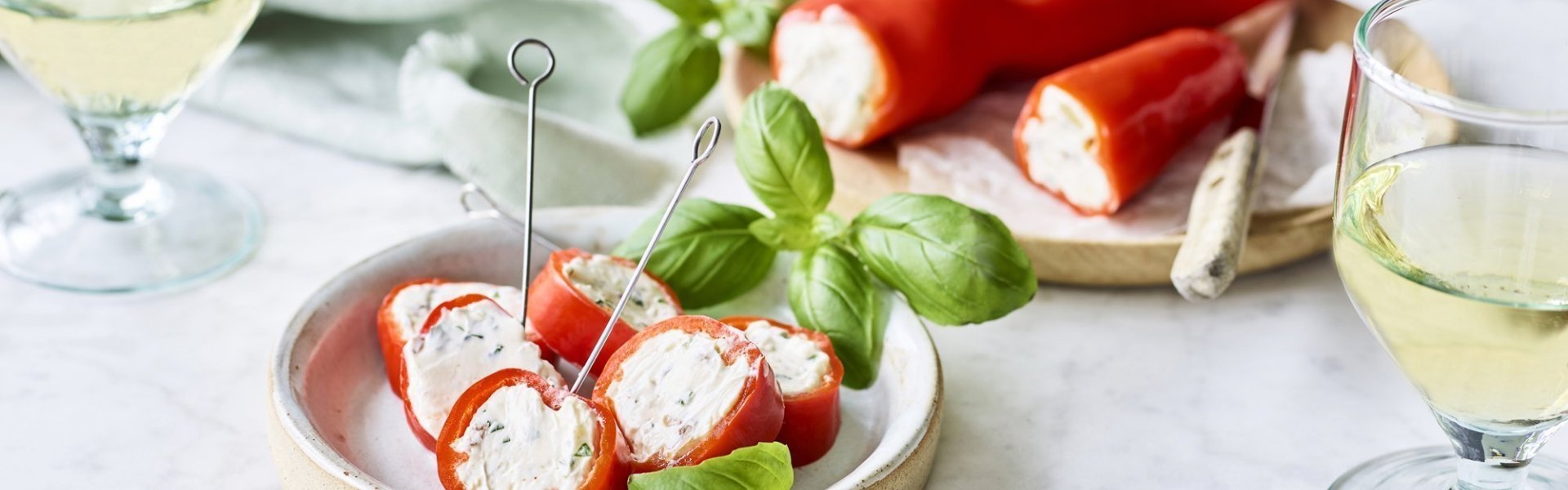 Snackpaprika met homemade kruidenkaas Een opgevulde puntpaprika met roomkaas en kruiden ligt op een houten snijplankje. Er werden rolletjes van gesneden en vastgeprikt met een stokje om het aperitief in stijl te starten. Voor de garnering en de kleur voegde we nog enkele verse blaadjes basilicum toe.