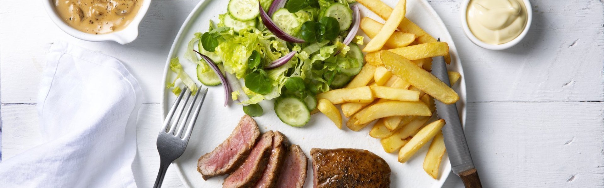 Een echte Vlaamse klassieker die smaakt voor jong & oud, een steak mooi gebakken met heerlijke goude frietjes en een salade. De champignonsaus staat langs het bord in een sauspotje. Bestek en servette liggen langs het bord klaar om te starten. 