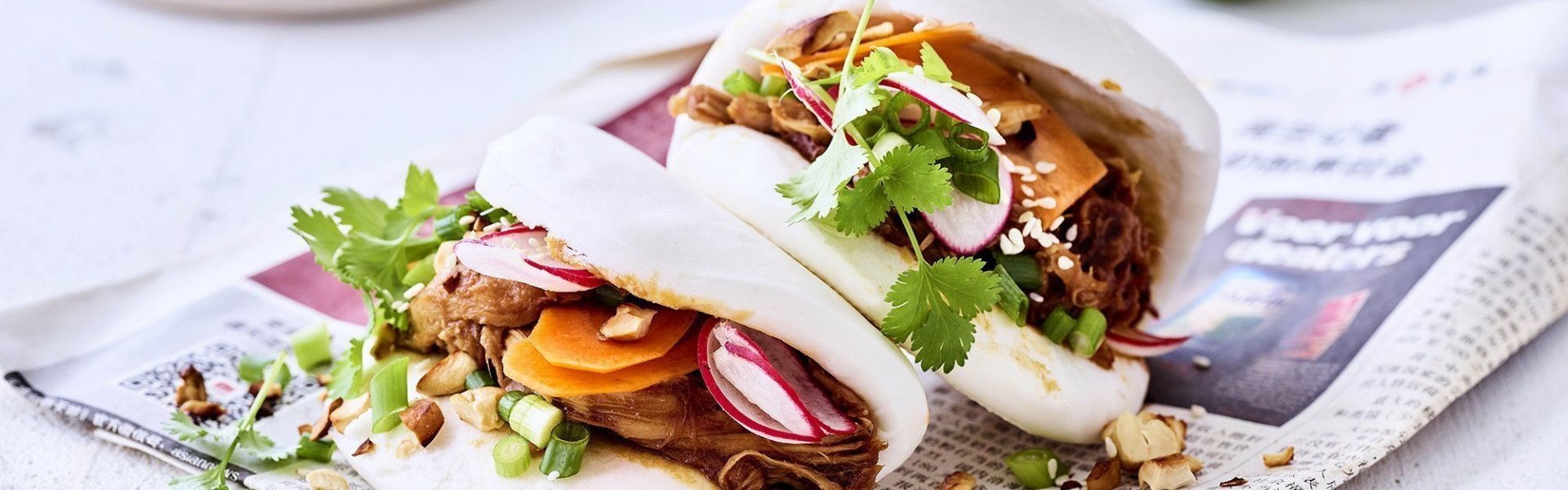 Steamed buns met pulled pork Steamed buns gevuld met pulled pork en knapperige groentjes zoals wortels en radijzen. Ze liggen op een krant op tafel met stukjes limoen erbij.