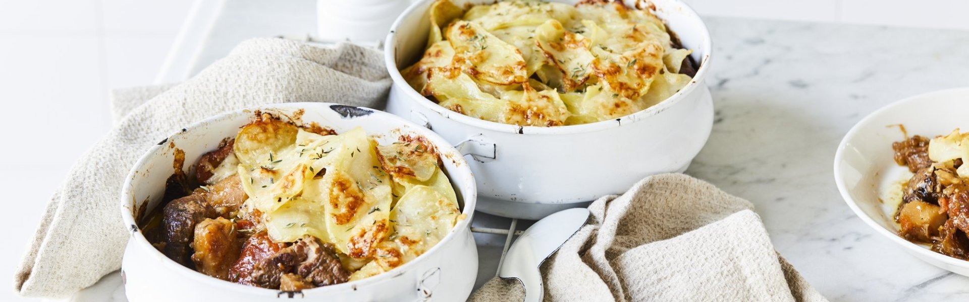 Twee witte stoofpotjes sttaan op een witte houten tafel en zijn gevuld met traag gegaard rundsvlees. De stoofpot is afgedekt met een laagje van aardappelschijfjes. De gratin is mooi goudgeel gebakken. Er staat een pepermolen langsheen de kookpotten, servette en grote lepel.