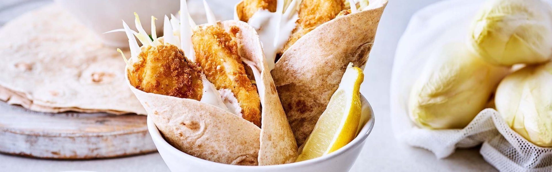 Taco's met gepaneerde visreepjes en een frisse witloofsalade Taco's met gepaneerde visreepjes en een witloofslaatje. De taco's zijn opgerold en zitten per 2 in een kommetje met een schijfje citroen. Het dipsausje staat apart naas de taco's voor wie extra wil.