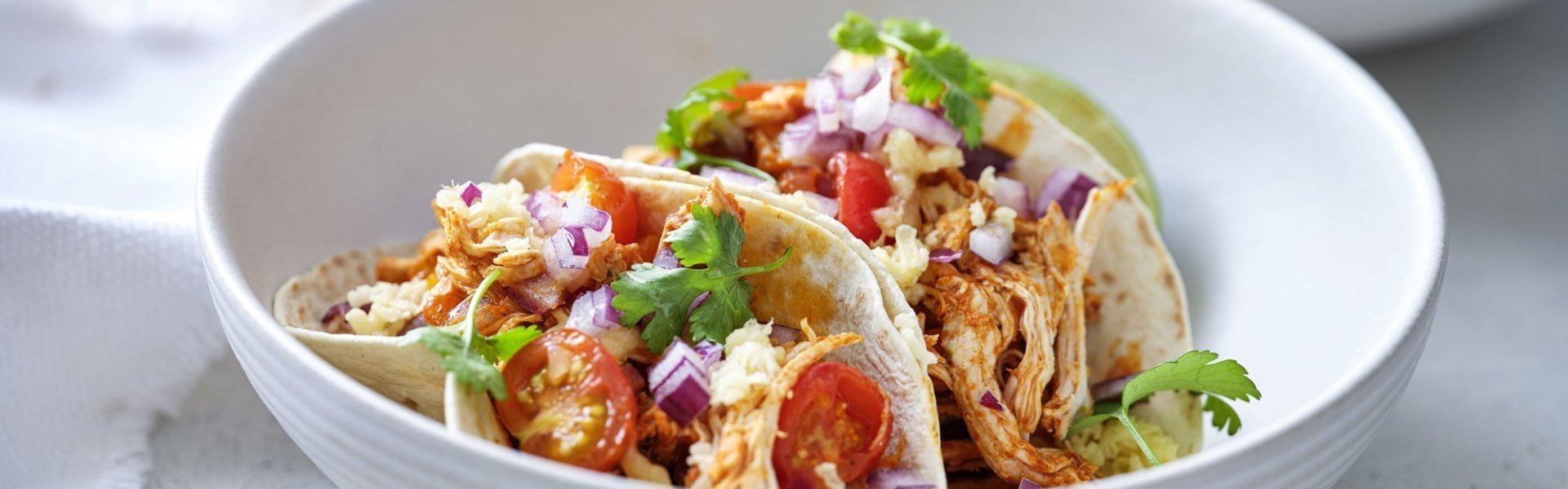 Taco’s met pulled chicken, tomaatjes, rode ui en Passendale kaas In een diep wit bord liggen twee gevulde taco's met pulled chicken, afgewerkt met tomaat, rode ui en geraspte Passendael kaas. Om wat extra kleur te geven liggen er enkele blaadjes koriander op. Wij serveren ze met frisgekoeld biertje, smakelijk!