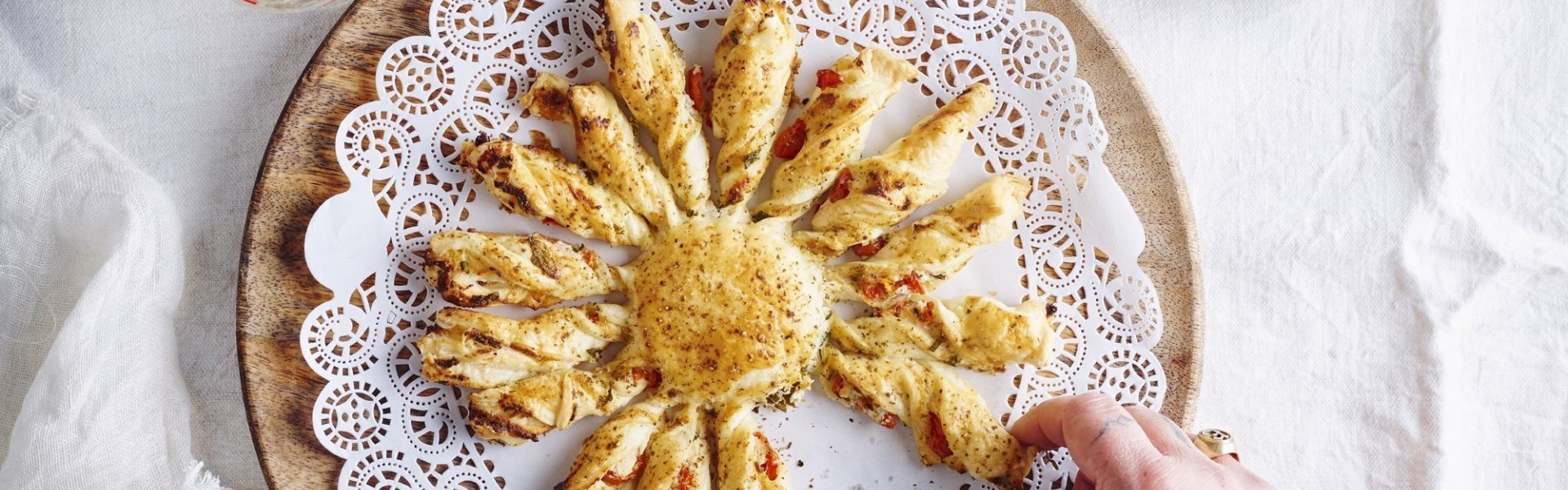 Tarte soleil met ricotta van berloumi en kerstomaat Een bladerdeeghapje met ricottakaas van bij ons en kerstomaatjes. Dit hapje is speciaal door de vorm, hij is nl gemaakt in de vorm van een zon. Hij ligt op een houten dienblad en er neemt een hand een zonnestraal om op te proeven.