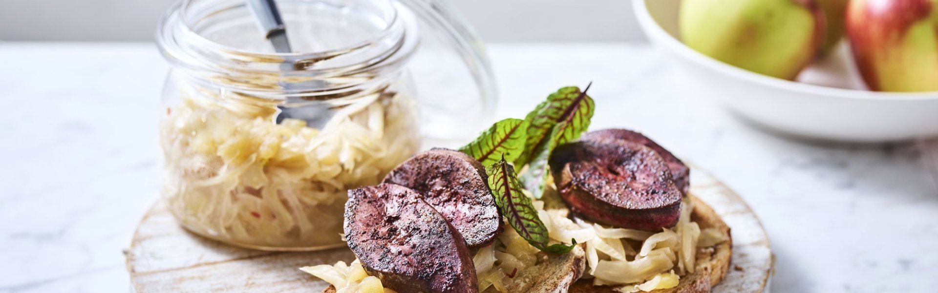 Toast met chutney van appel en venkel met gebakken varkensniertjes Twee toasten liggen op een houten serveerplank. Ze zijn belegt met chutney en gebakken niertjes. De overige chutney staat er langs in een glazen bokaal.